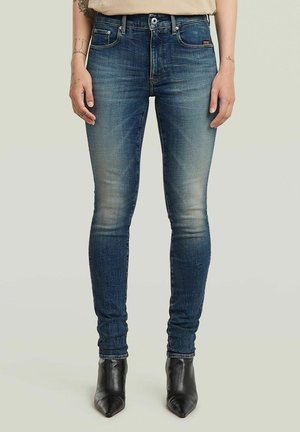 Jean slim fit en denim bleu avec un effet délavé, porté avec un haut beige et des bottines noires à bout pointu.