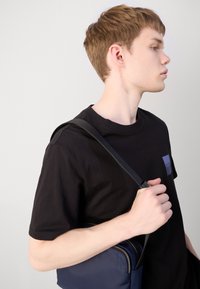 T-shirt en coton noir avec un petit logo carré bleu, porté avec un sac bandoulière marine présentant une fermeture éclair et une bandoulière texturée.