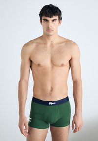 Green cotton boxer briefs with a navy waistband. Features a logo on the front. Smooth texture with a fitted design. Model stands relaxed.