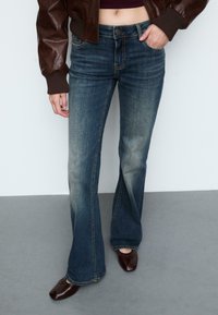 Rozšírené modré džínsy s vyblednutou textúrou, dizajnom s piatimi vreckami a kontrastným prešívaním. Skombinované s tmavohnedými topánkami.