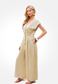 Beige wijde jumpsuit met een V-hals, korte mouwen, knoopsluiting aan de voorkant, een ceintuur in de taille en zakken. Draag het met chunky armbanden en sandalen.
