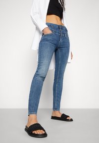 Mujer que lleva unos pantalones ajustados de cintura alta en azul con cierre de triple botón, un top negro corto, una camisa larga blanca y unas sandalias trenzadas negras con slide.