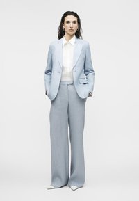 Femme debout, les mains dans les poches, portant un costume bleu clair, une chemise blanche et des chaussures blanches à bouts pointus, sur un fond gris uni.