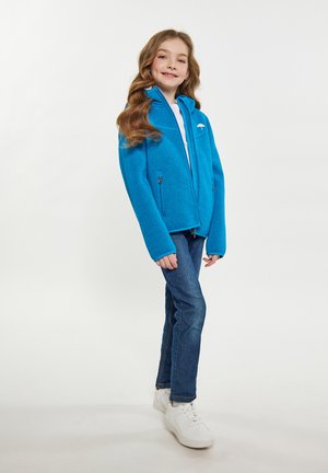 Giacca in pile blu brillante con colletto alto, due tasche con zip e polsini a contrasto, indossata sopra una maglietta bianca e jeans.