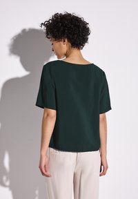 Tmavě zelený top s krátkými rukávy a širokým výstřihem, hladký na omak a volného střihu, v kombinaci se světlými kalhotami.