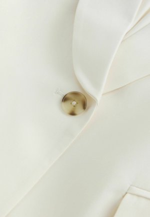 Ein einzelner brauner Knopf an einem cremefarbenen Kleidungsstück mit sichtbarem Revers und Taschenkante befestigt.