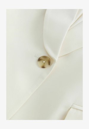 Ein einzelner brauner Knopf an einem cremefarbenen Kleidungsstück mit sichtbarem Revers und Taschenkante befestigt.