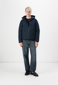 Navy blue puffer jacket with a hood, featuring quilted sections, zip closure, and elastic cuffs, paired with straight-leg denim jeans.