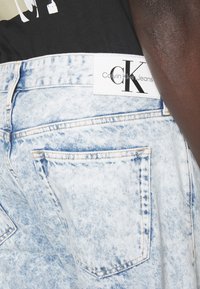 Los pantalones cortos de denim azul desgastado presentan una etiqueta blanca de Calvin Klein, cinco bolsillos y detalles de costura visibles en las costuras.