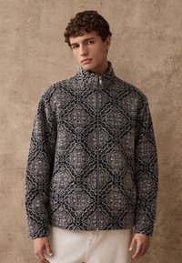 Fliisist jakk, millel on geomeetriline tumelilla ja helehall muster, püstkrae, kogu esiosa ulatuses lukuga ja pikad varrukad.