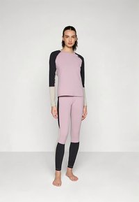 Žena koja stoji bosa, obučena u dug rukav majicu u ružičastoj, crnoj i bež boji te odgovarajuće tajice, nasuprot jednostavne bijele pozadine.