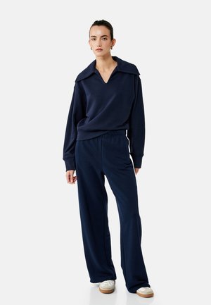 Ung kvinde iført navyblå langærmet top med bred krave og matchende vidde bukser, stående med afslappede hænder mod hvid baggrund.