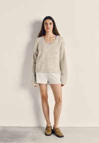 Bēšs mīksts adīts džemperis ar V veida kakla izgriezumu, pelēkie šorti un brūnas slip-on kurpes; mīksta tekstūra, brīvs piegriezums un garas piedurknes.