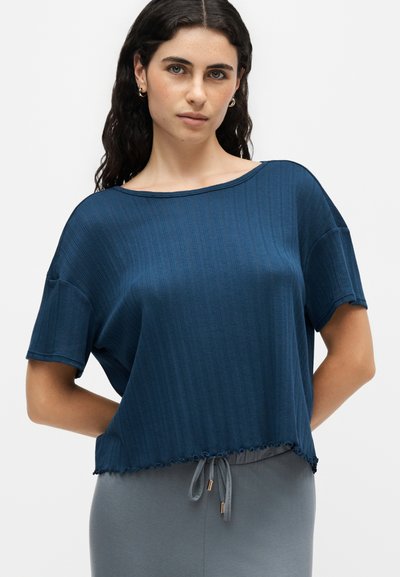 Femme aux longs cheveux foncés portant un haut bleu ample à manches courtes et un pantalon gris avec cordon, debout devant un fond uni.