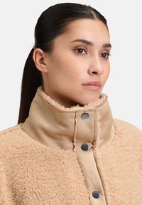 Veste en polaire beige avec un col montant ; fermeture par boutons ; texture douce ; matériau de col lisse contrastant.