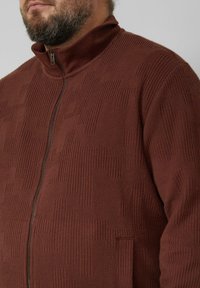 Brązowa kurtka na suwak z teksturowanym wzorem, z wysokim kołnierzem, przednimi kieszeniami i prążkowaniem. Wykonana z tkaniny tkanej.