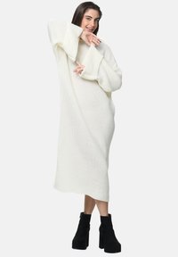 Gebreide witte oversized jurk met wijde mouwen, textuurstof en een losse pasvorm, gecombineerd met zwarte chunky-heeled enkellaarsjes.