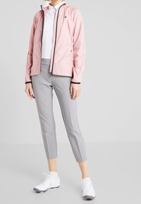 Hellrosa Reißverschlussjacke mit schwarzen Akzenten, graue eng anliegende Hose und weiße Sportschuhe. Verfügt über ein sportliches Design und feuchtigkeitsableitendes Material.