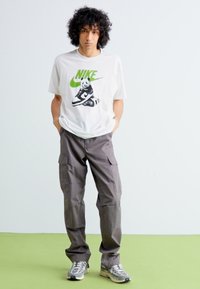 Hvid Nike t-shirt med grønt logo og tegneserie panda grafik, parret med grå cargo bukser og grå sneakers, sat op imod en lys baggrund.