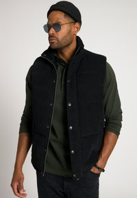 Schwarze Cordweste mit Druckknöpfen, hohem Kragen und Reißverschluss vorne. Getragen über einem dunkelgrünen Polohemd. Accessoires umfassen runde Sonnenbrillen.