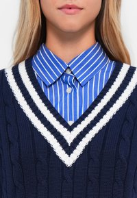 Frau, die ein blau-weiß gestreiftes Hemd mit Kragen unter einem marineblauen, kabelgestrickten Pullover mit weißen und marineblauen V-Streifen trägt.