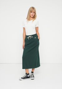 Grønn maxi skjørt i myk stoff med snøring i livet, kombinert med en tettsittende hvit t-skjorte med korte ermer og svarte høye sneakers.