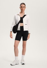 Fehér cipzáras dzseki fekete kiegészítőkkel, fekete crop top-pal és hozzá illő, magasított derékú kerékpáros rövidnadrággal párosítva. Fekete átvetős táska, fehér sportcipő.