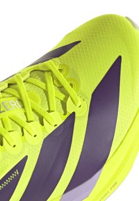 Primo piano di una scarpa da corsa gialla neon con dettagli a zig zag neri, lacci dettagliati e tomaia in tessuto leggero e testurizzato.