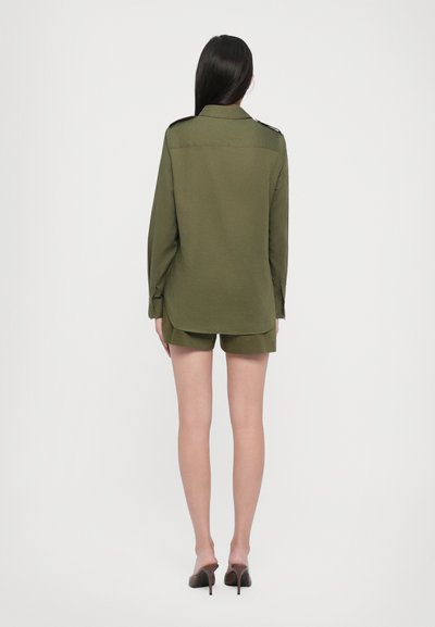 Olivovo zelená košeľa s dlhým rukávom a detailmi na pleciach, spárovaná s rovnakými šortkami a čiernymi vysokými podpätkami. Hladká textúra, voľný strih.