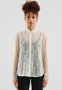 Witte kanten blouse met doorschijnende lange mouwen en een kraag, met een knoopsluiting aan de voorkant en een bloemig kanten patroon, gecombineerd met zwarte broek.