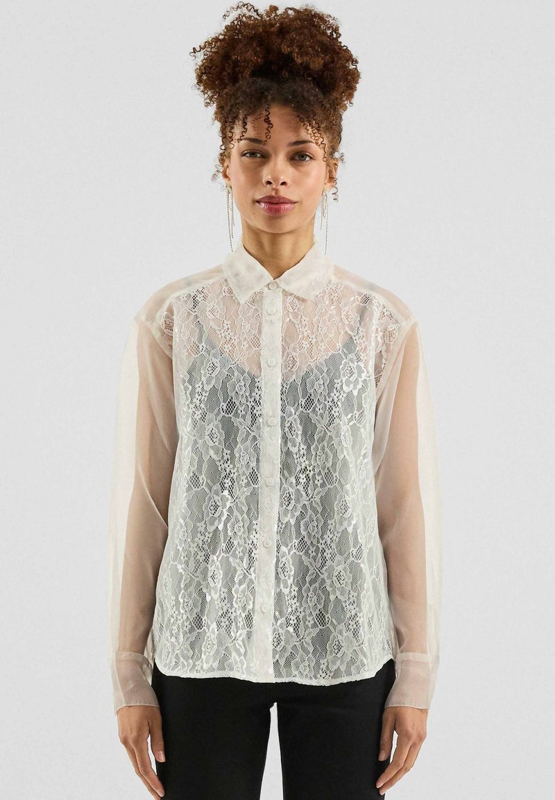Witte kanten blouse met doorschijnende lange mouwen en een kraag, met een knoopsluiting aan de voorkant en een bloemig kanten patroon, gecombineerd met zwarte broek.