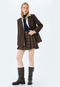 Bruine getailleerde blazer over een marineblauwe top en een geruite minirok. Slate grijze kniehoge laarzen met gespen en roomkleurige sokken.
