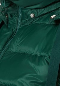 Zielona, pikowana kamizelka z wysokim kołnierzem, błyszczącą teksturą, zamek błyskawiczny i zatrzaski oraz regulowane paski na ramionach.