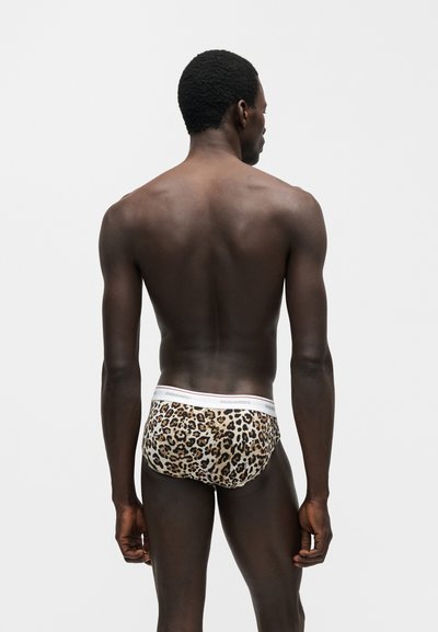 Άντρας με την πλάτη γυρισμένη, φοράει εσώρουχο με λεοπάρ σχέδιο και λευκή ζώνη μέσης, στέκεται μπροστά σε απλό ανοιχτό γκρι φόντο.