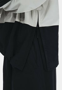 Gros plan d'une veste noire et beige avec une fermeture éclair latérale et un détail en tissu superposé sur un pantalon noir.