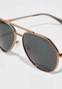 Óculos de sol em metal com armação bronze, lentes octogonais e tonalidade cinza. Apresenta uma ponte dupla e almofadas de nariz transparentes para conforto.