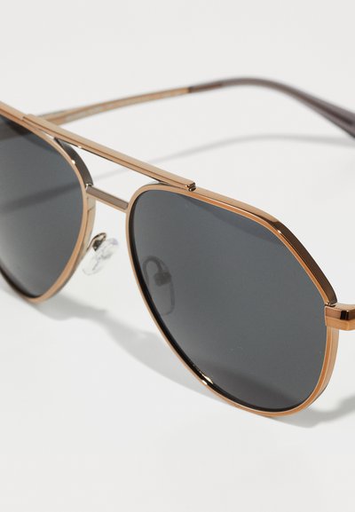 Lunettes de soleil en métal avec une monture en bronze, des verres octogonaux et une teinte grise. Présentent un double pont et des plaquettes de nez transparentes pour plus de confort.