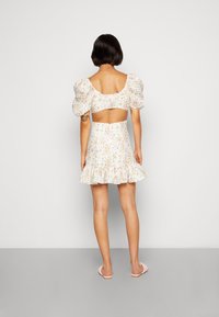 Robe fleurie avec manches bouffantes, fond blanc, ourlet à volants, dos ouvert et taille accentuée, assortie à des sandales rose clair.