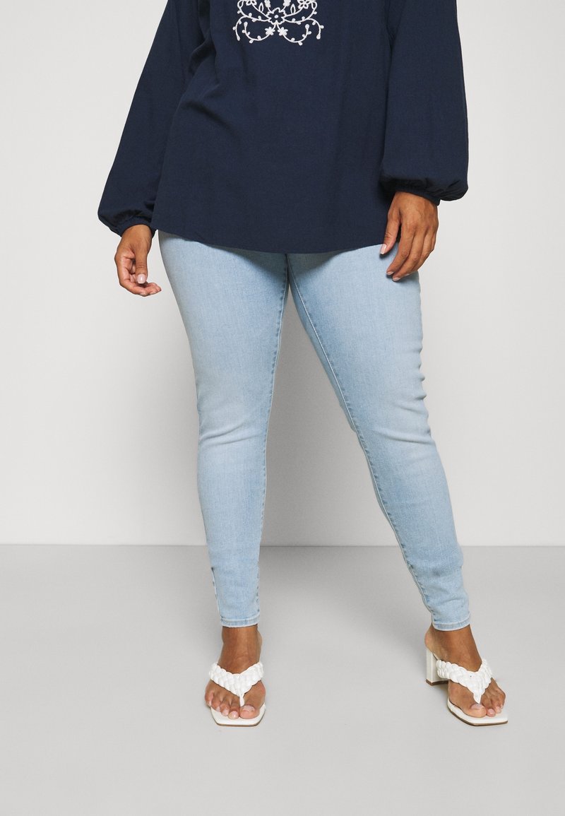 Personne portant un jean skinny bleu clair, un chemisier bleu marine avec une broderie blanche et des sandales blanches à talons, debout devant un fond uni.