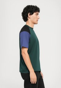 Giovane uomo con capelli ricci che indossa una maglietta verde con maniche bloccate in nero e blu, in posa di profilo su uno sfondo semplice.