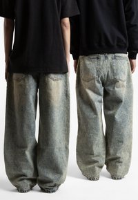 Baggy jeans iz denima v svetlo modrem pranju, z elastičnimi manšeti, vidnimi šivi in dvema zadnjima žepoma, v kombinaciji s črnimi majicami.