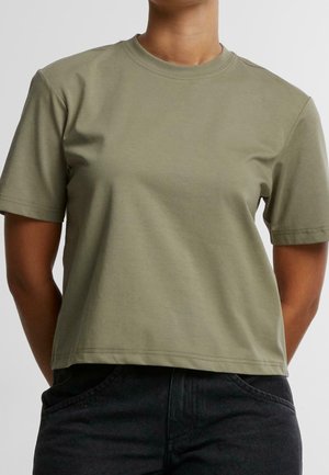 Person trägt ein schlichtes, kurzärmliges, olivgrünes Cropped-T-Shirt und dunkle, hoch taillierte Jeans und steht vor einem einfarbigen Hintergrund.