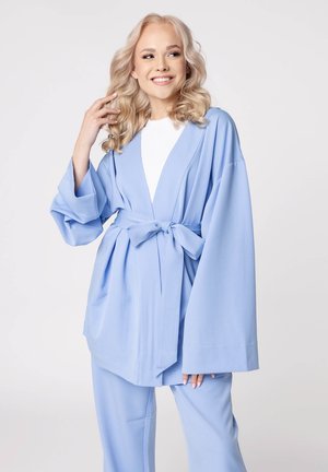 Veste bleu clair avec une ceinture à nouer, manches larges et coupe décontractée, associée à un pantalon assorti et un haut blanc en dessous. Texture de tissu douce.