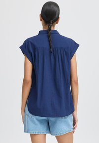 Camicia blu navy a maniche corte con colletto a bottoni, caratterizzata da una texture a costine verticali e dettagli arricciati sul retro, abbinata a pantaloni shorts in denim.