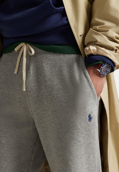 Pantaloni de trening gri din bumbac cu un cordon de ajustare la talie, având un logo albastru pe coapsa stângă. Sunt purtați cu un hanorac bleumarin și o cămașă verde.