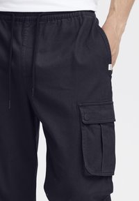 Pantaloni cargo negri, confecționați dintr-un material moale, cu un tiv elastic, având buzunare laterale și un buzunar mare cu clapetă pe coapsă.