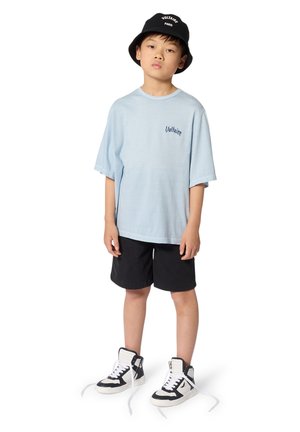 Lichtblauw oversized T-shirt met "Voltaire" logo, gecombineerd met zwarte shorts en zwart-witte hoge sneakers met witte veters.