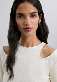 Haut blanc cassé en tissu texturé, avec un design à épaules dénudées et un col arrondi. Cheveux lisses et foncés partiellement visibles.