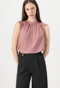Roosa ilma varrukat blouse, millel on pitsist õlavarre aktsendid, koos kogutud kaeluse ja võtmeauguga. Kombineeritud kõrge piha mustade pükstega.