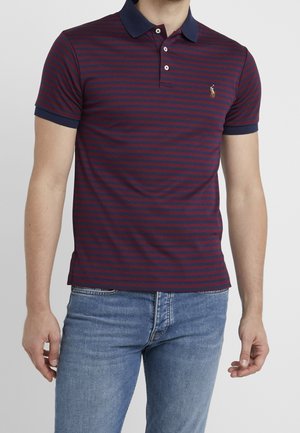 Mężczyzna w granatowo-bordowej koszulce polo w paski z kołnierzykiem i jasnoniebieskich jeansach, stojący na jednolitym tle.